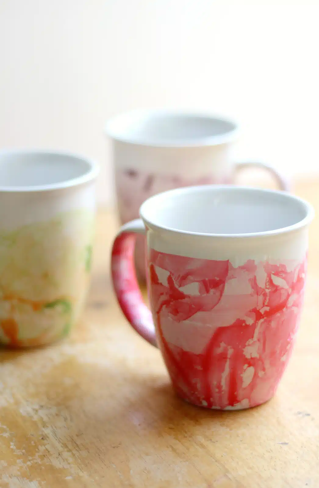 Three ceramic mugs with marbled designs in red, green, and purple rest on a wooden surface, perfect examples of crafts for kids to make and sell.