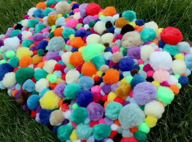 A colorful assortment of pom-poms of various sizes and colors is spread on a grassy surface, perfect for inspiring crafts for kids to make and sell.