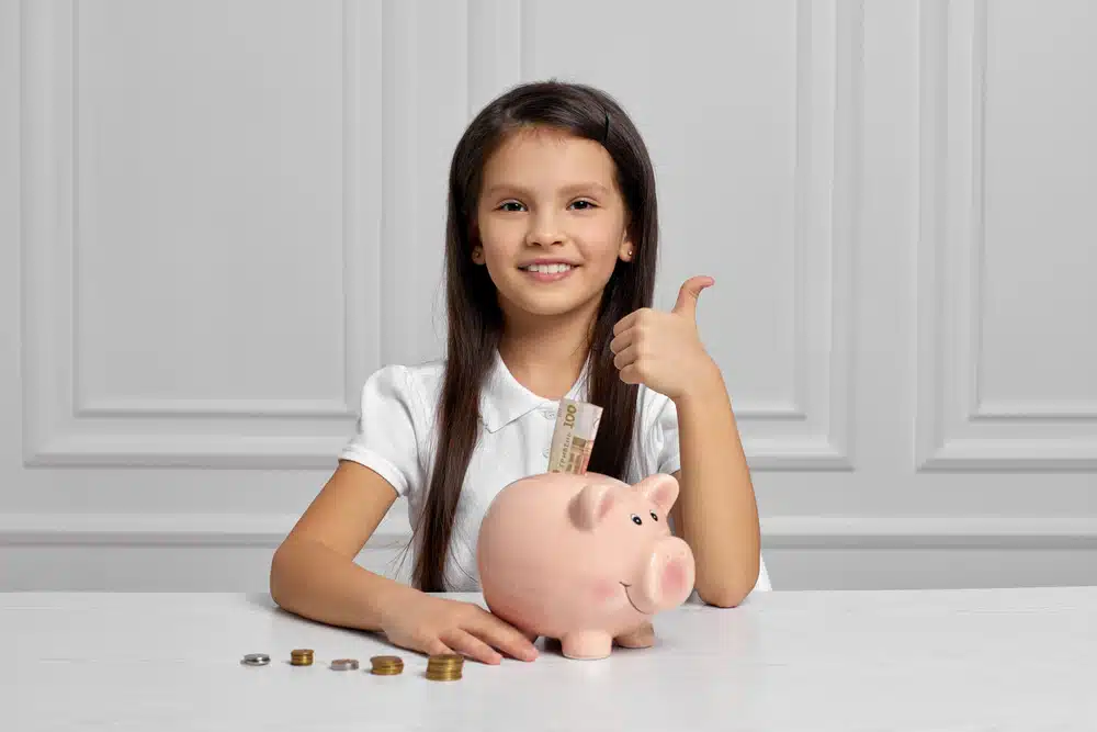 A girl in a white shirt sits at a table with a pink piggy bank, giving a thumbs-up amidst coins and a banknote, perhaps planning smart strategies for things to sell at school.