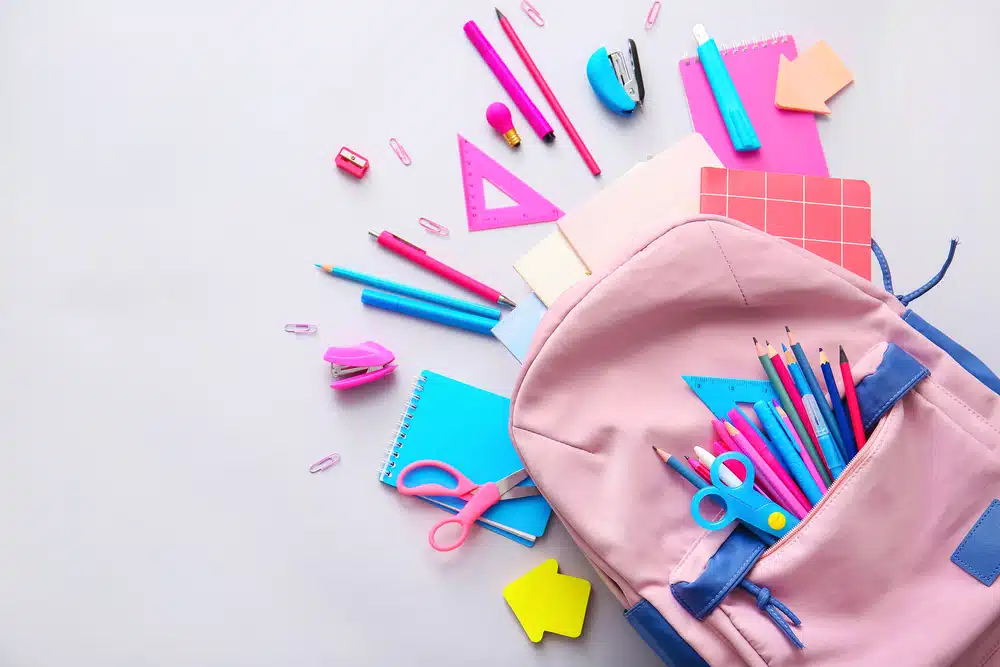 Open pink backpack with assorted school supplies, like notebooks, pencils, scissors, and erasers—perfect things to sell at school—scattered on a light gray background.