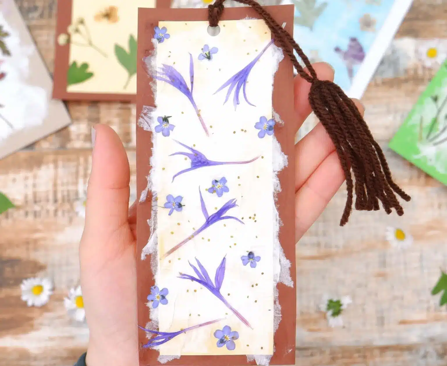 A hand holds a handmade bookmark with pressed purple flowers and petals on a brown and white background, featuring a brown tassel. These charming crafts for kids to make and sell can also be seen in the background.