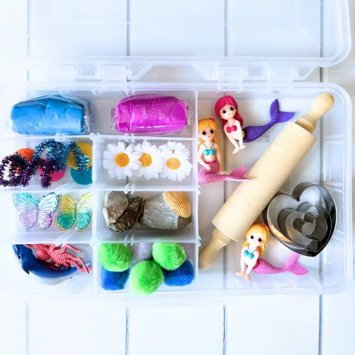 Explore the clear plastic organizer filled with playdough, plastic mermaid figures, rolling pin, heart-shaped cutters, pom poms, daisies, butterflies, rocks, and toy dinosaurs—perfect for inspiring crafts for kids to make and sell.