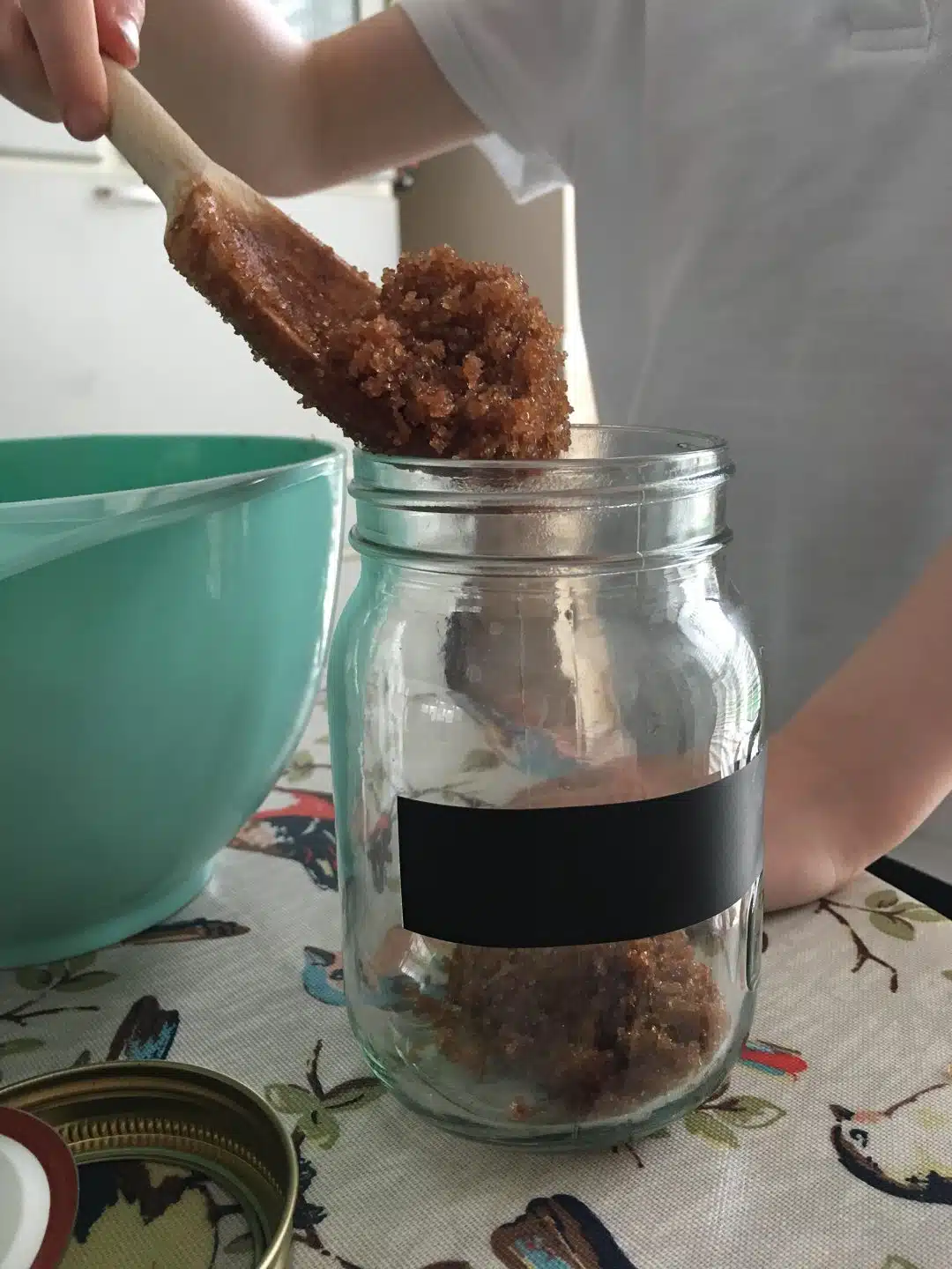 A child in a white shirt carefully spoons a brown sugar mixture from a green bowl into a glass jar on a patterned tablecloth, creating charming DIY gifts perfect for crafts for kids to make and sell.