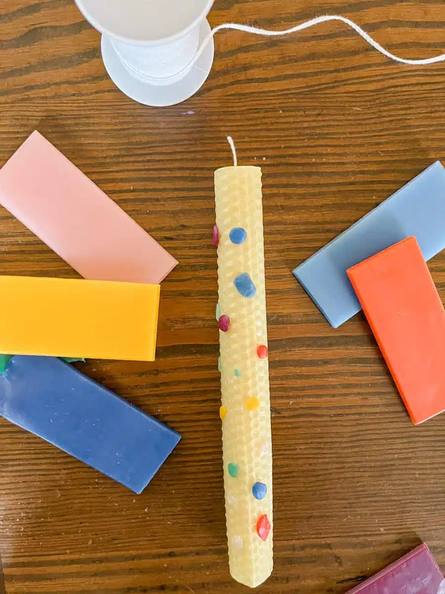 A rolled beeswax candle, perfect for crafts for kids to make and sell, is decorated with colorful wax bits. It lies on a wooden table, surrounded by vibrant wax blocks and a spool of white wick, ready to spark creativity.
