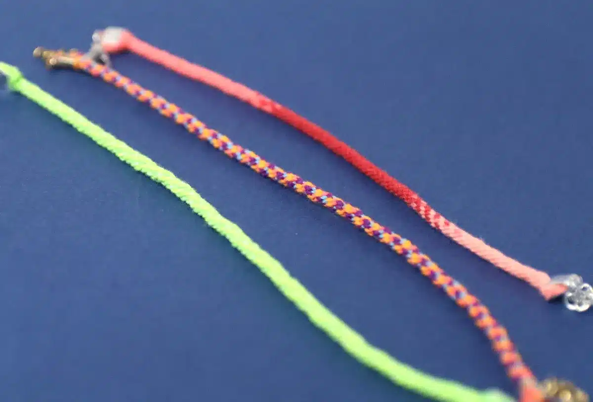 Three braided friendship bracelets, perfect crafts for kids to make and sell, are laid out on a blue surface. The vibrant colors include pink, a multicolored mix of orange, blue, and purple, and neon green.